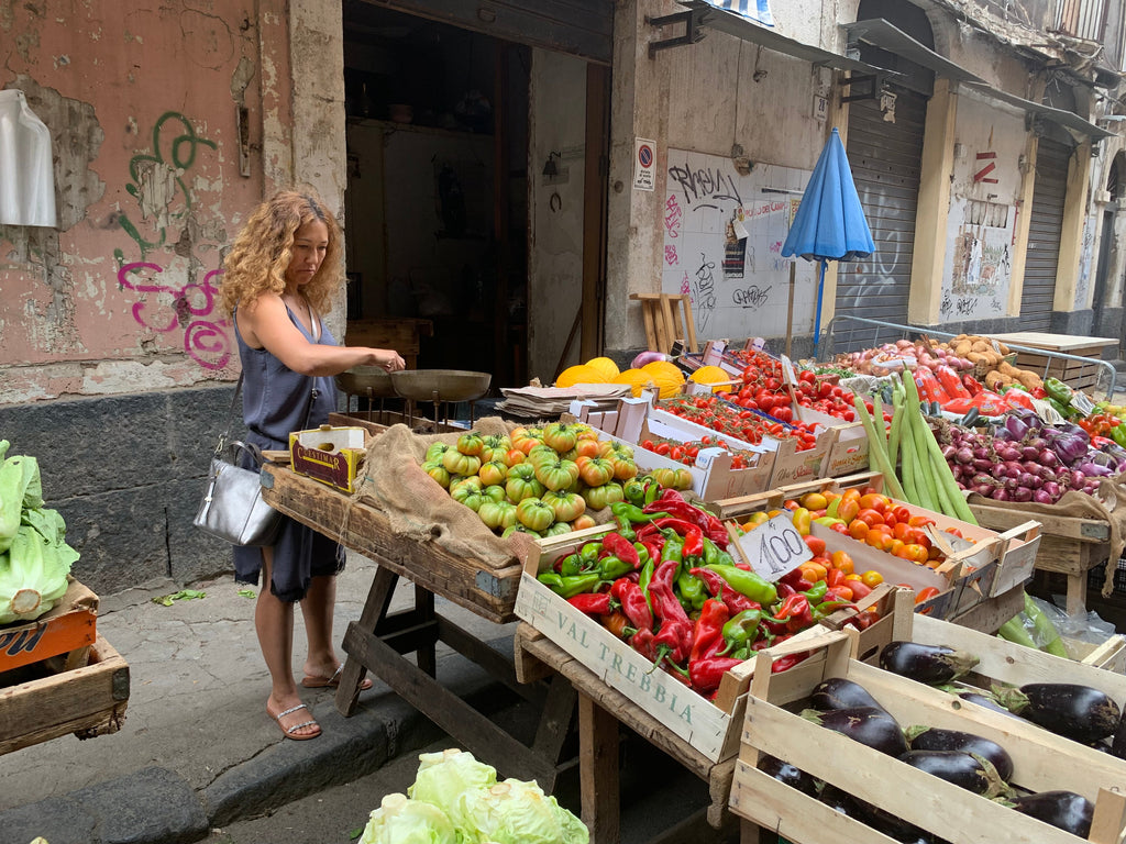 A tourist explores a Sicilian outdoor market with colorful produce stands and people walking along narrow streets